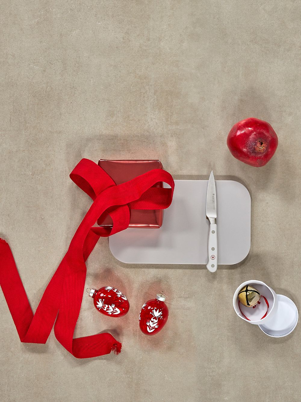 Red gift box with ribbon, knife, and pomegranate on a beige surface