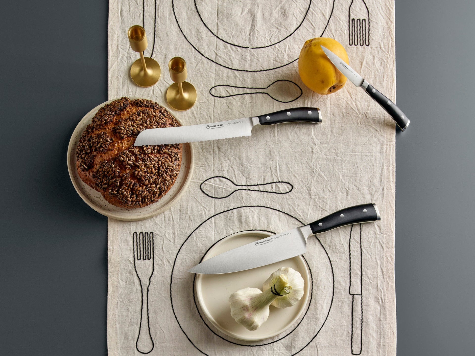 Table setting with knives on a table runner, featuring a seeded loaf, garlic, a pear, and brass candle holders.