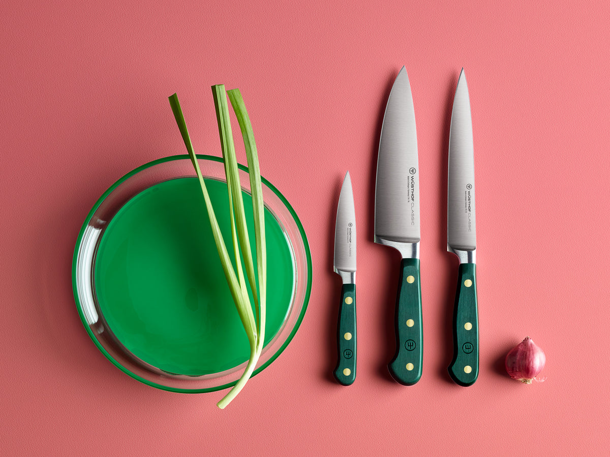 Three knives with green handles on a pink background, next to a green plate with green onions and a small red onion.