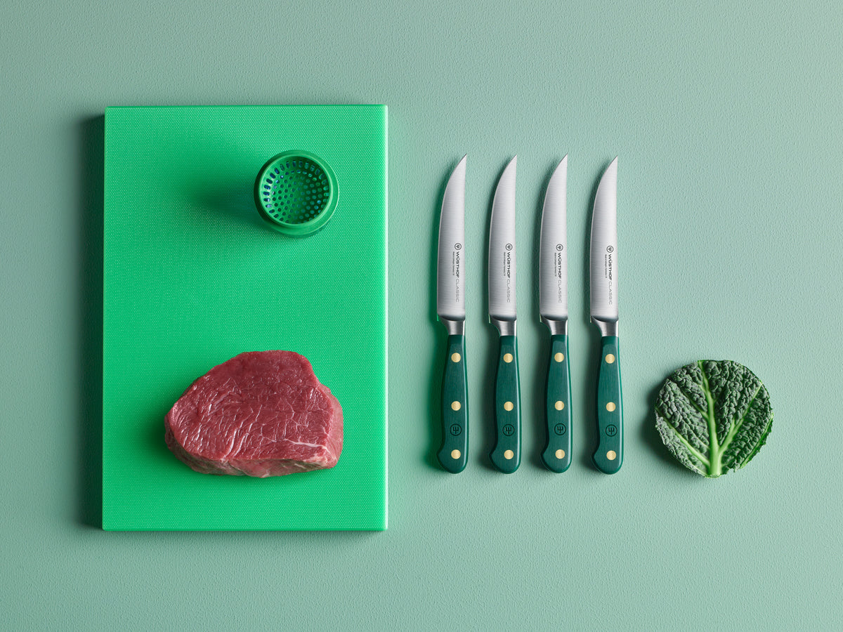 Green cutting board with raw meat, a green kitchen tool, and four knives on a light green background