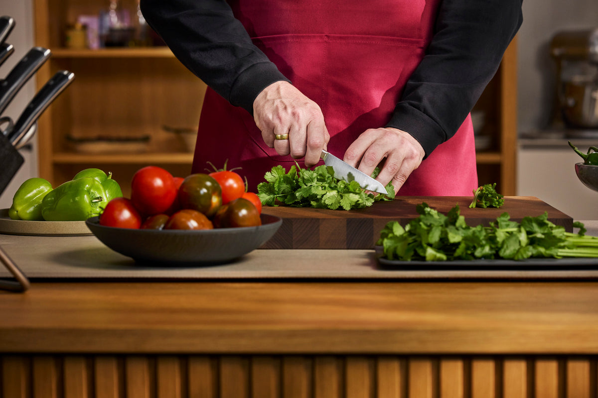 A person wearing a pink apron over a black long-sleeve shirt chops fresh green herbs on a wooden cutting board in a kitchen setting. A bowl of red and green tomatoes and a tray of chopped parsley are visible on the counter.