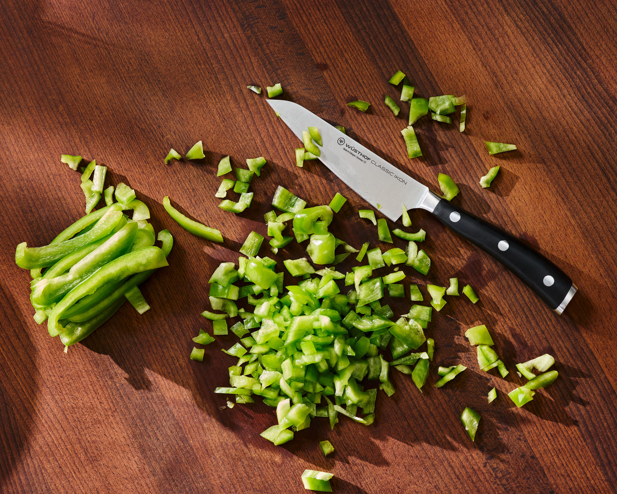 A Victorinox kitchen knife with a black handle and silver blade rests on a dark wooden cutting board next to chopped green bell peppers and a pile of sliced green peppers.