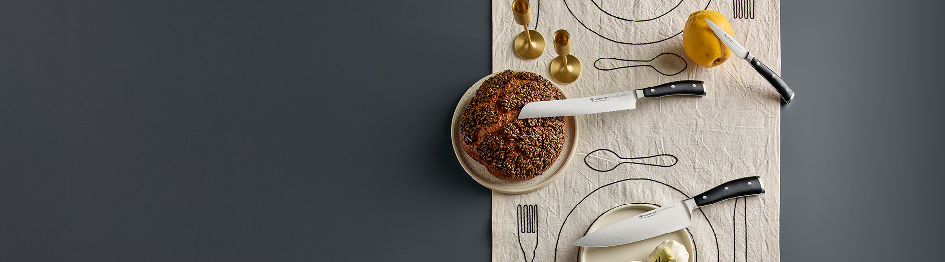 A table setting with knives, a loaf of bread, a pear, and gold measuring spoons on a patterned tablecloth.