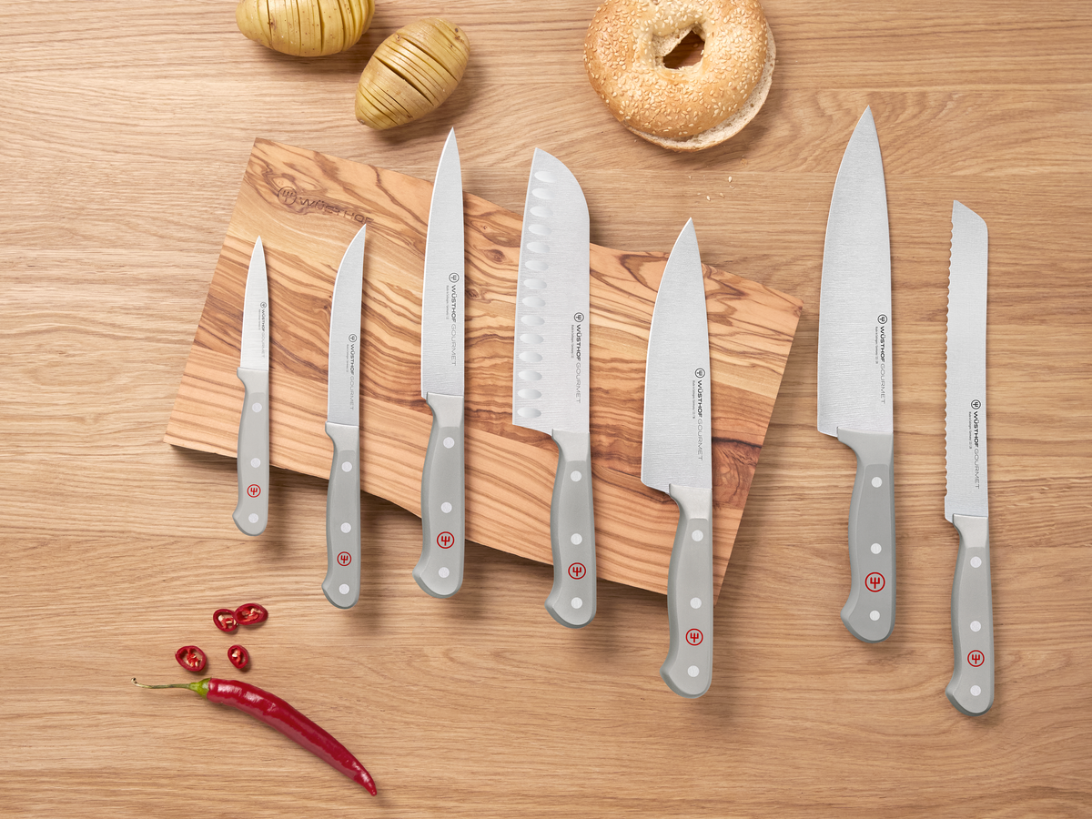 Set of knives on a wooden cutting board with bread and vegetables on a wooden surface