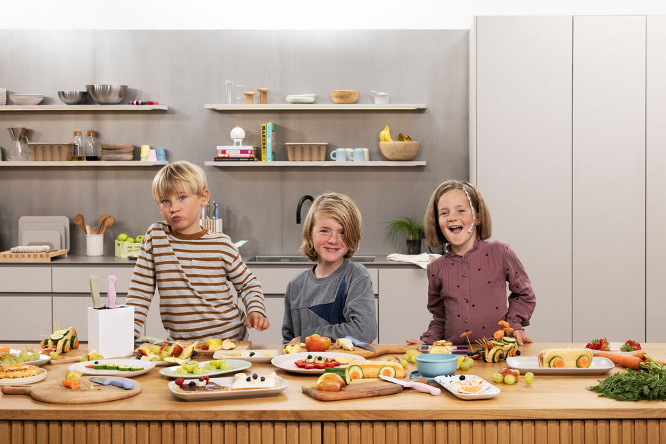 Three children in a kitchen with food on plates and vegetables on a counter.