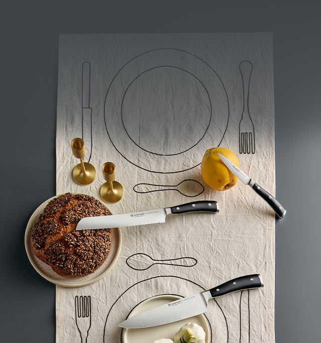 Knife set on a table with a bread loaf, a sliced apple, and gold candle holders on a tablecloth with plate and cutlery outlines.