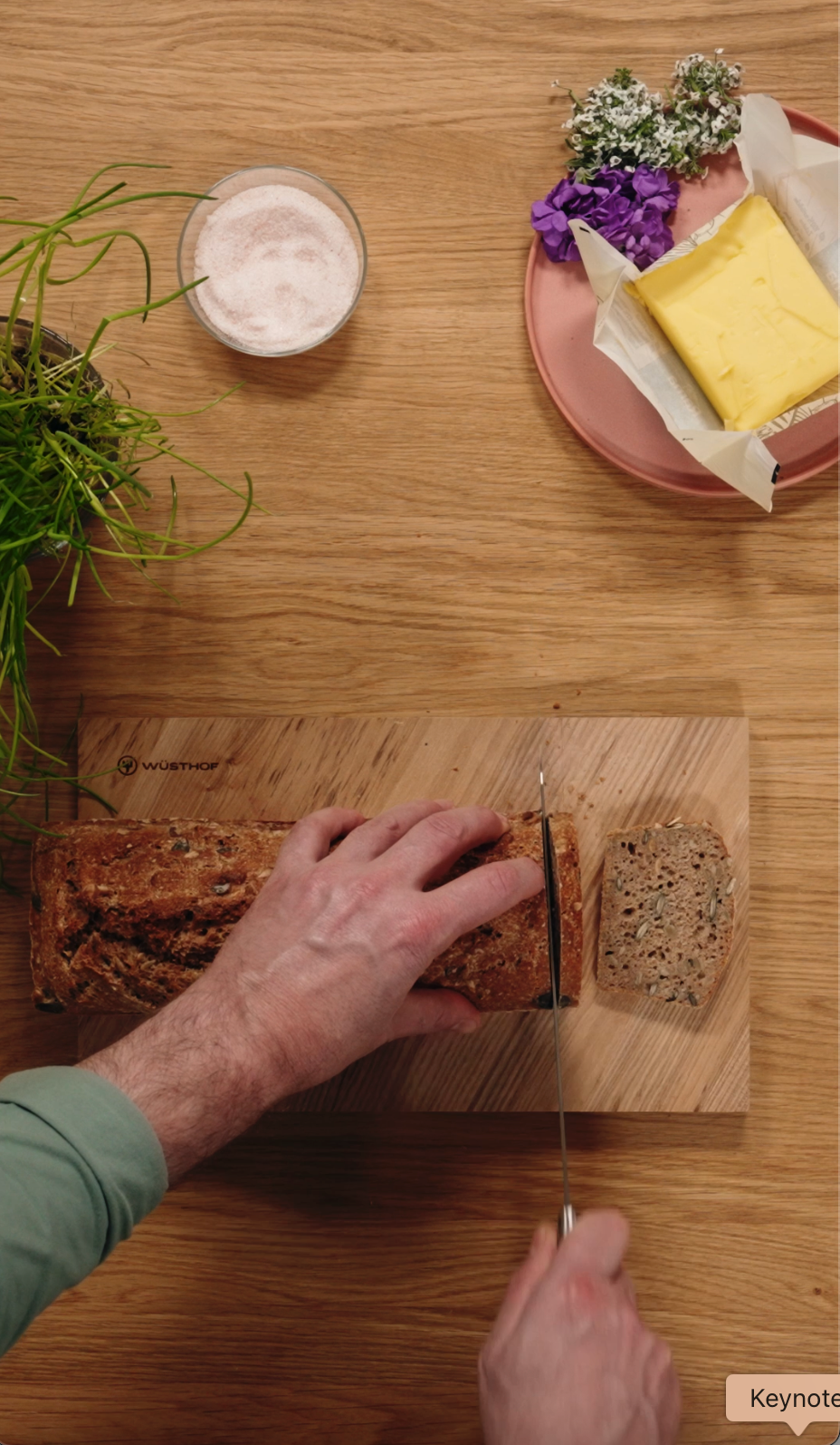 Person slicing bread on a wooden cutting board with butter and flowers in the background.