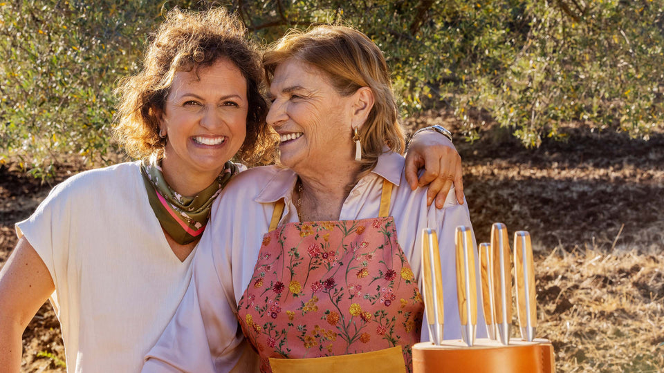 Two women standing outdoors with a rustic background, one wearing an apron.