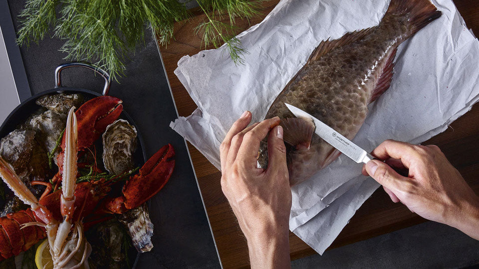 Person preparing a fish with a knife on a wooden surface, surrounded by other seafood items.