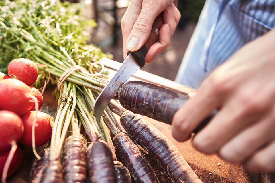 Person cutting purple carrots with a knife on a wooden board, surrounded by radishes.