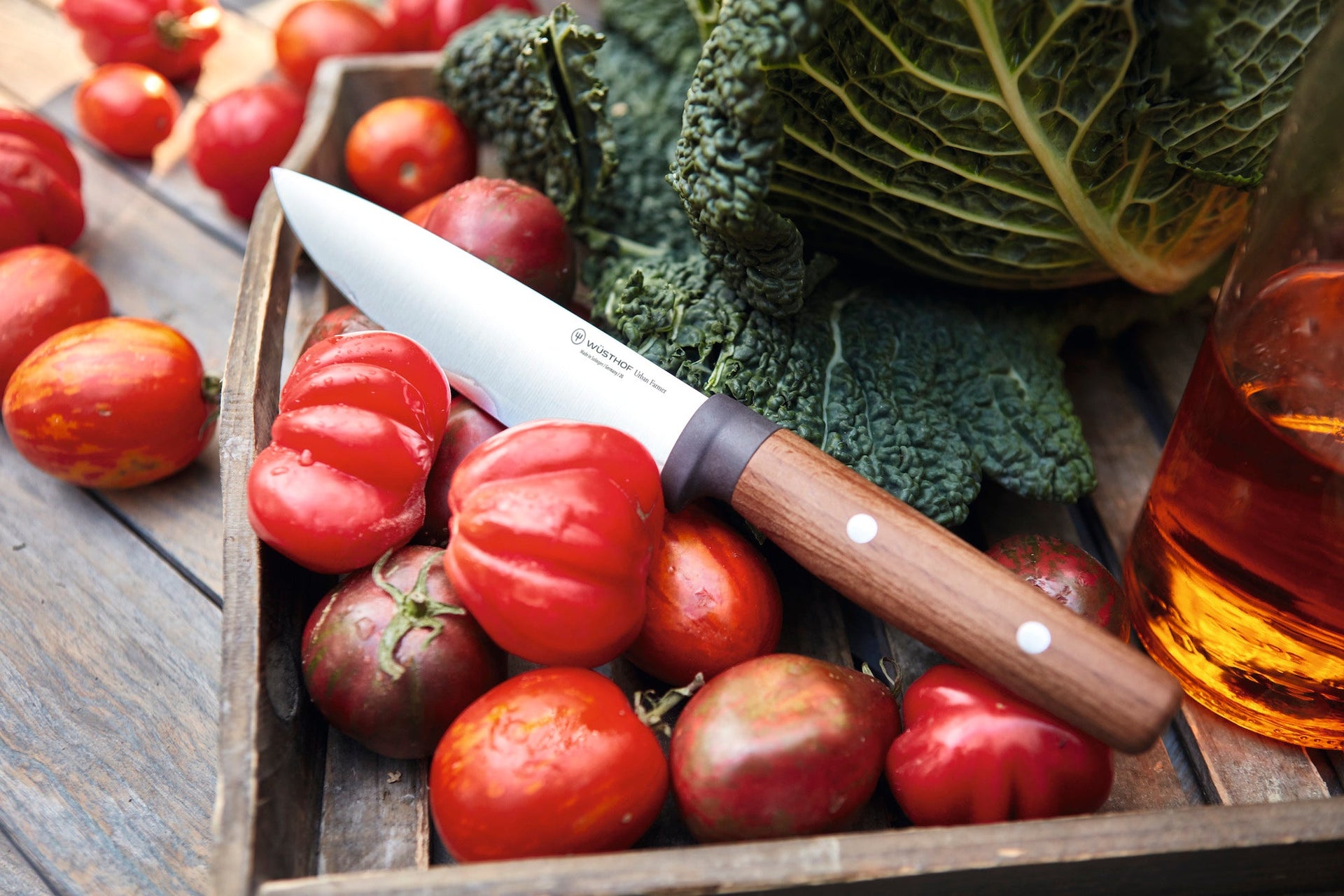 Tomatoes in a wooden box with a knife, kale, and a bottle of oil on a rustic wooden surface.