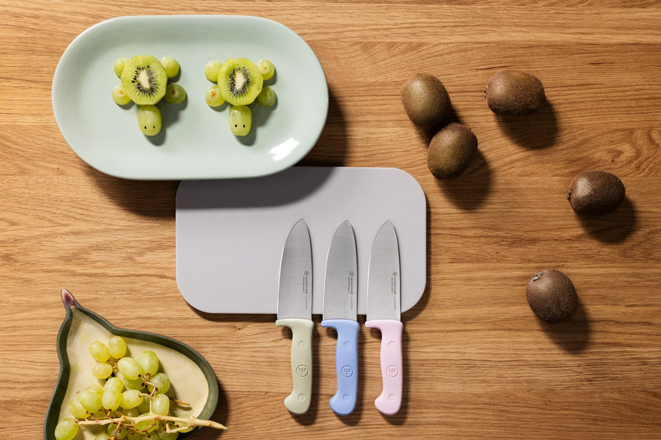 Children's knife set on cutting board, sitting on a wooden table surrounded by kiwis