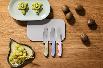 Three knives with colored handles on a wooden surface next to kiwis and grapes.