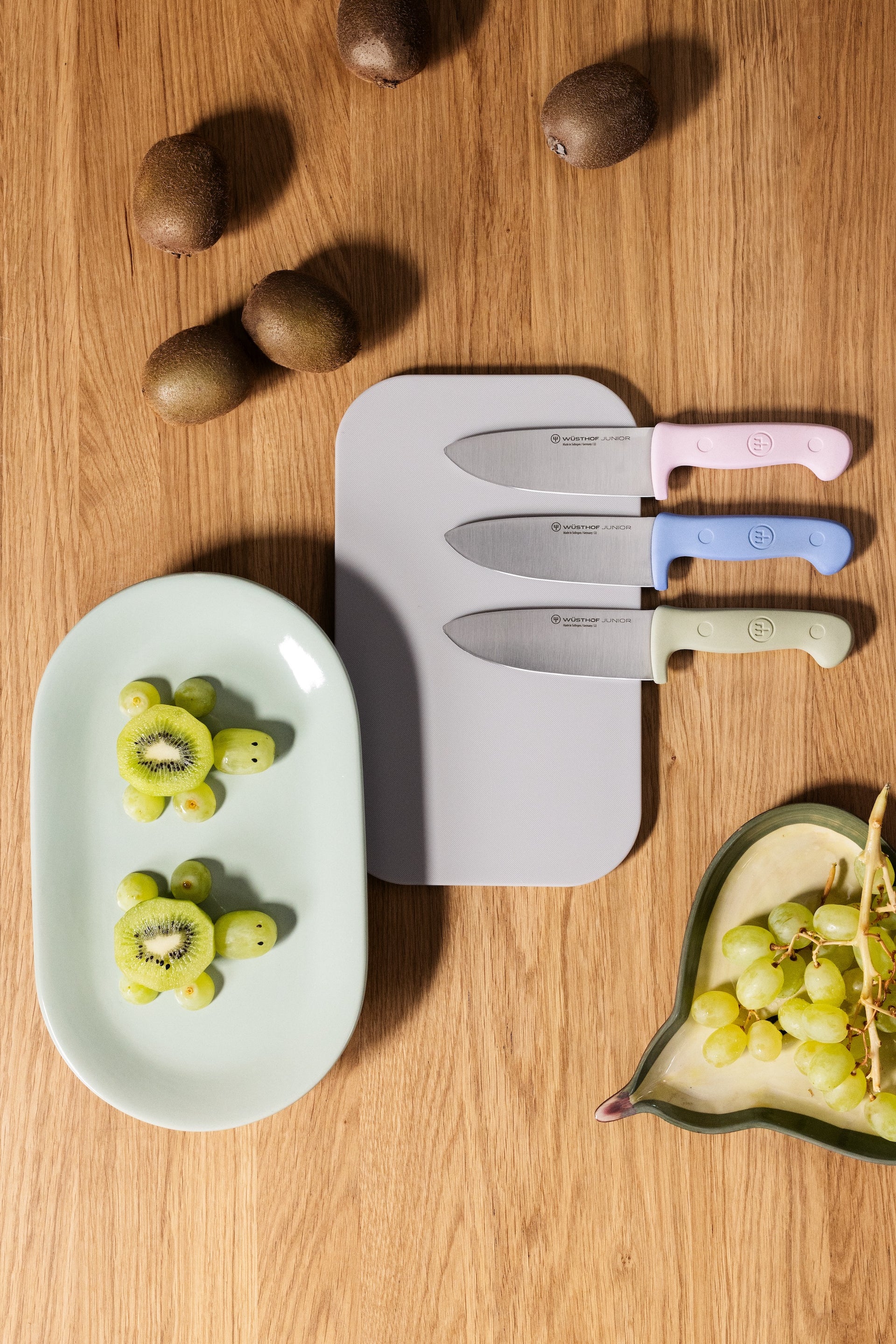 Children's knife set on cutting board, sitting on a wooden table surrounded by kiwis