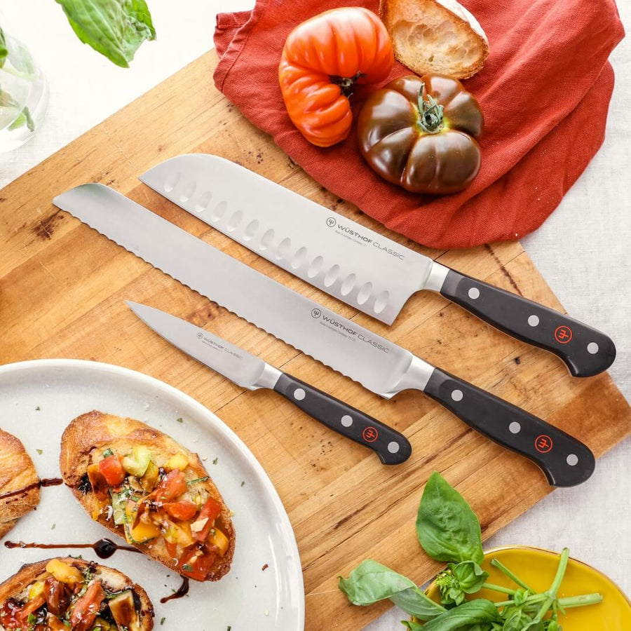 Three knives on a wooden cutting board with vegetables and a plate of food.